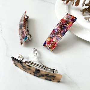 May include: Three hair clips with different patterns. One clip is brown with black spots, one is brown with blue and pink spots, and one is purple with pink and white spots.