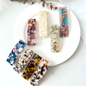 May include: Assortment of colorful, patterned hair clips. The clips are made of a translucent plastic material and have a rectangular shape. The patterns include abstract designs, floral motifs, and animal prints.