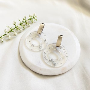 May include: A pair of clear resin hoop earrings with silver flecks and silver metal hooks.