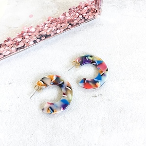 May include: A pair of colorful, multi-colored, hoop earrings. The earrings are made of a white, translucent material with swirls of blue, red, yellow, and purple.