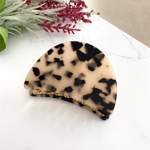 May include: A half-moon shaped tortoiseshell hair clip with a black and white pattern. The clip has a gold-colored metal hinge.