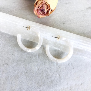 Könnte beinhalten: Ein Paar weiße, halbkreisförmige Creolen mit Marmoreffekt. Die Ohrringe haben goldfarbene Stifte und werden auf einer weißen, strukturierten Oberfläche mit getrockneten Rosen im Hintergrund präsentiert.