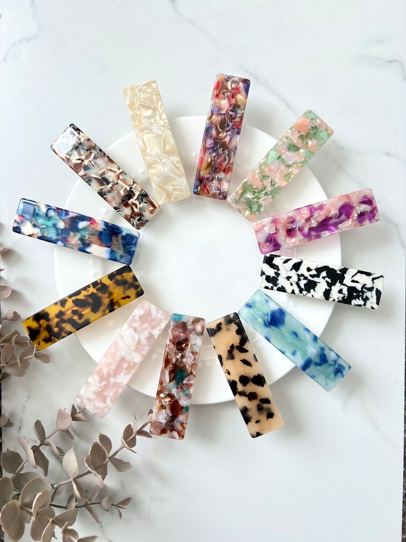 May include: Assortment of twelve colorful rectangular hair clips with various patterns, including tortoise shell, leopard print, and abstract designs. The clips are arranged in a circle on a white plate.
