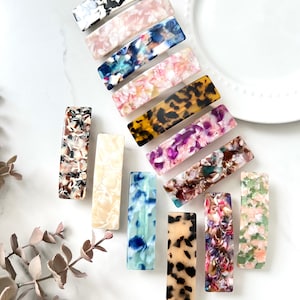 May include: Assortment of 12 colorful patterned hair clips. The clips are made of a translucent plastic material with a variety of patterns, including leopard print, floral, and abstract designs. The clips are arranged on a white surface.