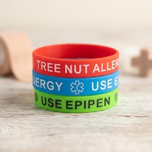 May include: A stack of three silicone medical alert wristbands. The top band is red with white text reading TREE NUT ALLERGY. The middle band is blue with white text and a medical symbol. The bottom band is green with black text reading USE EPIPEN.