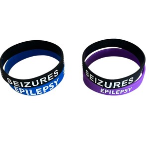 K&ouml;nnte beinhalten: Zwei schwarze Silikonarmb&auml;nder mit wei&szlig;em Text. Das obere Armband hat ein blaues Innenband und lautet "SEIZURES EPILEPSY". Das untere Armband hat ein violettes Innenband und lautet "SEIZURES EPILEPSY".