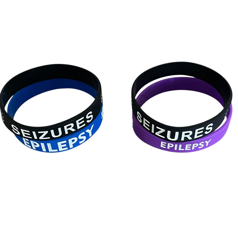 K&ouml;nnte beinhalten: Zwei schwarze Silikonarmb&auml;nder mit wei&szlig;em Text. Das obere Armband hat ein blaues Innenband und lautet "SEIZURES EPILEPSY". Das untere Armband hat ein violettes Innenband und lautet "SEIZURES EPILEPSY".