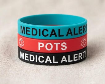 Puede incluir: Una pila de tres pulseras de silicona. La banda superior es de color verde azulado con texto negro que dice MEDICAL ALERT. La banda central es roja con texto blanco que dice POTS y dos símbolos de estrella médica. La banda inferior es negra con texto blanco MEDICAL ALERT!