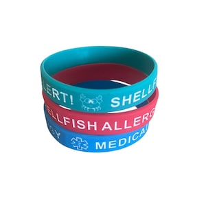 May include: Three stacked silicone wristbands in teal, red, and blue. The teal band reads "SHELLFISH ALERT!" with a crab graphic. The red band says "SHELLFISH ALLERGY". The blue band says "MEDICAL ALERT" with a medical symbol.