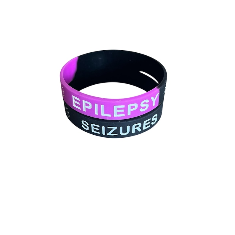 K&ouml;nnte beinhalten: Zwei schwarze Silikonarmb&auml;nder mit wei&szlig;em Text. Das obere Armband ist pink und sagt "EPILEPSIE". Das untere Armband sagt "ANF&Auml;LLE".