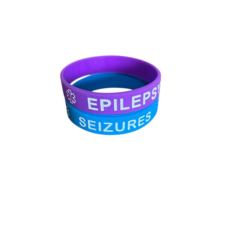 K&ouml;nnte beinhalten: Zwei Silikonarmb&auml;nder, eines lila und eines blau. Das lila Armband hat ein wei&szlig;es Stern-des-Lebens-Symbol und den Text "EPILEPSIE". Das blaue Armband hat den Text "ANF&Auml;LLE".