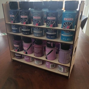 May include: A wooden paint bottle organizer with two shelves. The organizer holds 12 bottles of acrylic paint in various shades of blue and purple. The bottles are labeled "Craft Acrylic" and are 2 fl oz (59 ml).