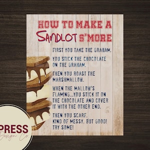May include: A wooden sign with the text "HOW TO MAKE A SANDLOT S'MORE" in red and blue. The sign includes instructions and an illustration of a s'more. The Express Design Co. logo is in the bottom left corner.