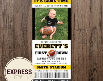 May include: A yellow and black football-themed first birthday party invitation with a photo of a baby boy holding a football. The invitation says "It's Game Time! Our little boy is turning one!" and "Please join us for Everett's First Down. Saturday, October 5. Kick Off - 11:00 a.m. Concession stand opens at half-time. Smith Stadium. Optional attire - favorite sports team. Sec 10, Row 05, Seat 19."