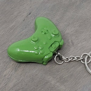 May include: A green plastic video game controller keyring with a silver chain and ring. The keyring is a replica of a popular gaming console controller.