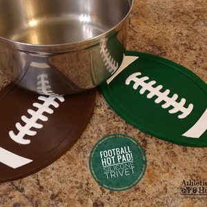 Pode incluir: Dois tapetes de silicone em forma de bola de futebol, um marrom e outro verde, com costuras brancas. O texto "Football Hot Pad! Silicone Trivet" está em um círculo verde.