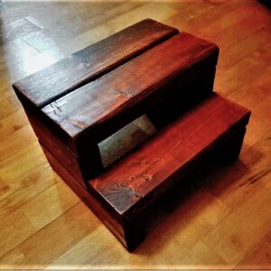 Puede incluir: Un taburete de madera de dos peldaños con un tinte marrón oscuro. El taburete está hecho de madera maciza y tiene un aspecto rústico.