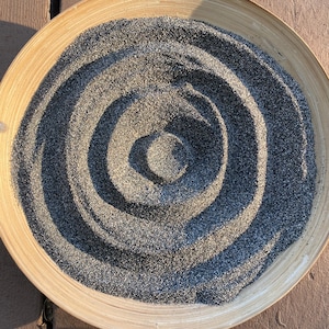 May include: A round, light-colored bamboo tray filled with dark gray sand arranged in concentric circles. The sand has a textured appearance, creating a pattern of shadows and highlights. The tray sits on a wooden surface.