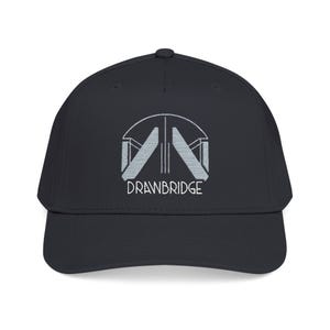 Peut inclure: Casquette de baseball noire avec une visière incurvée. Le chapeau présente un logo brodé blanc avec le mot "DRAWBRIDGE" dans une police stylisée. Le logo représente un design architectural abstrait.