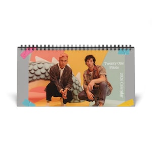 May include: A spiral-bound desk calendar featuring two people. The calendar has a gray border with the text "Twenty One Pilots 2026 Calendar". The background has a colorful design with a dragon-like sculpture.