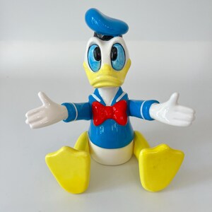 May include: A Donald Duck figurine with a blue sailor shirt, bow tie, and hat. The figure has a yellow beak and feet, white gloves, and is posed with arms outstretched. The cartoon character is a classic Disney collectible.