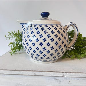 May include: A ceramic teapot with a white base and a blue floral pattern. The teapot has a rounded body, a curved handle, and a spout. The lid is topped with a blue knob. The teapot is sitting on a white surface.