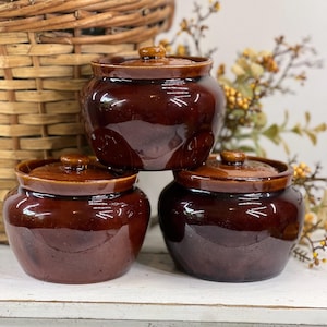 May include: Three small, round, dark brown glazed ceramic pots with matching lids. Each pot features a wide, bulbous body and a short, narrow neck with a flat rim. The glossy finish reflects light, highlighting the smooth, rounded shape of the vessels.
