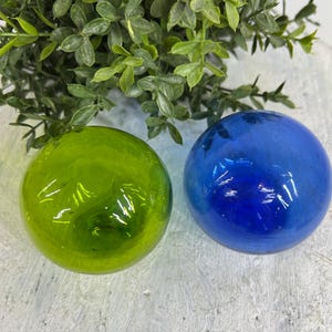 May include: Two glass spheres, one green and one blue, sit side-by-side. The green sphere has a glossy finish, while the blue sphere is a deep, saturated color. Green leafy foliage is in the background.