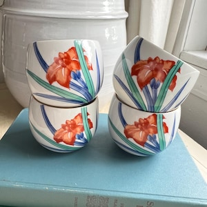 May include: Four small, white ceramic bowls stacked on a blue book. Each bowl features a vibrant orange flower design with green and blue accents. The bowls have a rounded shape and a gold rim, showcasing a floral theme.
