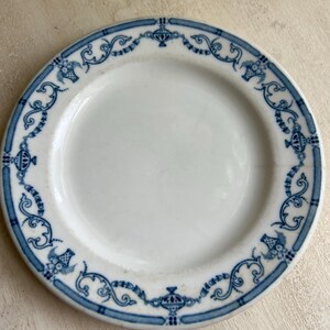 May include: White ceramic plate with a blue decorative border. The border features a repeating pattern of floral and scroll designs. The plate has a simple, elegant design, suitable for serving food or as a decorative piece.