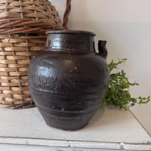 May include: A dark brown, antique-style ceramic jar with a small spout and a textured surface. The jar is set on a white surface, with a woven basket and greenery in the background. The jar has a rustic, aged appearance.