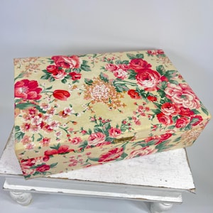 May include: A rectangular wooden box with a floral pattern featuring red and pink roses, green leaves, and small white flowers on a cream background. The box has a gold-colored clasp.