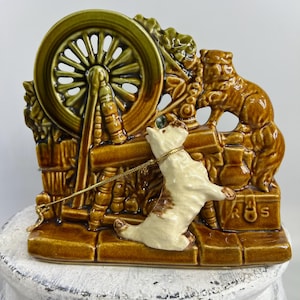 May include: A ceramic figurine depicting a dog pulling a cart with a spinning wheel. The figurine is primarily brown and cream-colored, with green accents on the wheel. The dog is on a gold chain. The figurine has a vintage aesthetic.