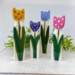 May include: Four ceramic tulip vases in yellow, blue, purple, and pink, each with a different polka dot pattern. The vases have green leaves and stems, and are set against a white background with greenery.