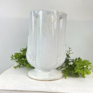 May include: A white ceramic vase with a pedestal base. The vase has a fluted design and a glossy finish. Green foliage is placed around the base of the vase. The vase is a decorative item.