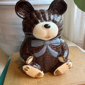 May include: A brown ceramic teddy bear figurine with a woven texture. The bear has a white face, tan paws, and a blue bow. The bear is sitting on a vintage book.