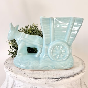 May include: A light blue ceramic donkey pulling a cart planter. The planter has a detailed design of a donkey, cart wheels, and a textured base. The planter is filled with green foliage. The planter is on a white, distressed pedestal.