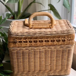 May include: A rectangular, light brown wicker basket with a woven handle and a decorative cross-stitch pattern around the lid. The basket is designed with a hinged lid and is suitable for storage or decorative purposes.