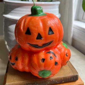 May include: A stack of three orange pumpkin-shaped candles with black triangle eyes and smiles, and green stems. The candles are sitting on top of a stack of old books with orange and brown covers. A white pot with a green plant is in the background.