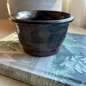 May include: A small, dark blue and brown ceramic pot with a wide rim. The pot has a speckled glaze and a band of brown around the middle.