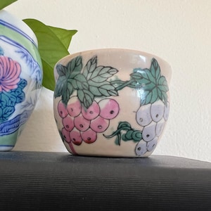 May include: A small, white ceramic planter with a pink and purple grape design. The planter has a round shape and a slightly flared lip.