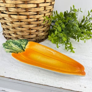May include: A ceramic serving dish shaped like a carrot with a bright orange body and textured green leafy top. The dish has a glossy finish and an elongated, tapered form, designed to hold small items or food, showcasing a realistic vegetable design.
