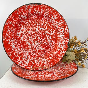 May include: Two round enamel plates featuring a vibrant red and white speckled splatter pattern. Each plate has a smooth, glossy finish and a thin black rim around the edge. The design consists of irregular red paint spots over a white base.
