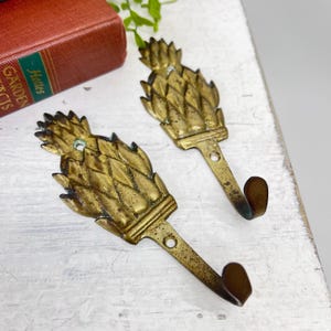 May include: Two antique brass wall hooks. Each hook features a detailed artichoke design at the top, with a curved hook below. The hooks are a warm, aged brass color, suggesting vintage or antique origins. They are likely used for hanging items.