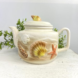 May include: A cream-colored ceramic teapot featuring a textured sand-like finish decorated with raised, three-dimensional sea life elements including a starfish, a scallop shell, a spiral shell, a sand dollar, and a small shell on the lid.