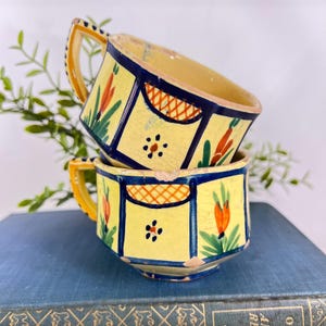 May include: Two stacked ceramic cups featuring a pale yellow glaze with dark blue geometric borders. The cups are decorated with hand-painted orange floral motifs and green leaves, accented by small orange cross-hatched patterns on the upper rim.