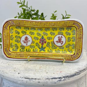 May include: A rectangular yellow ceramic dish with a red and gold border. The dish features Chinese characters and floral designs. The dish is supported by a gold-colored metal stand.