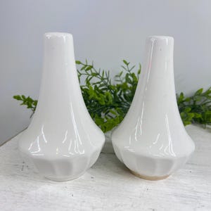 May include: Two white ceramic salt and pepper shakers. The shakers have a pear-shaped design with a flared base and a smooth, glossy finish. The base has a ridged texture. The shakers are displayed on a white wooden surface.