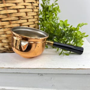 May include: A small copper saucepan with a polished metallic exterior and a silver-toned interior. The pot features a single pouring spout on the side and a long, straight black handle attached with a brass-colored metal ferrule at the base.