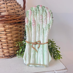 May include: A ceramic asparagus-shaped serving dish. The dish features a bundle of asparagus stalks in shades of white, green, and pink, tied with a gold-colored ribbon. The dish is supported by a gold metal stand, and is a decorative kitchen item.